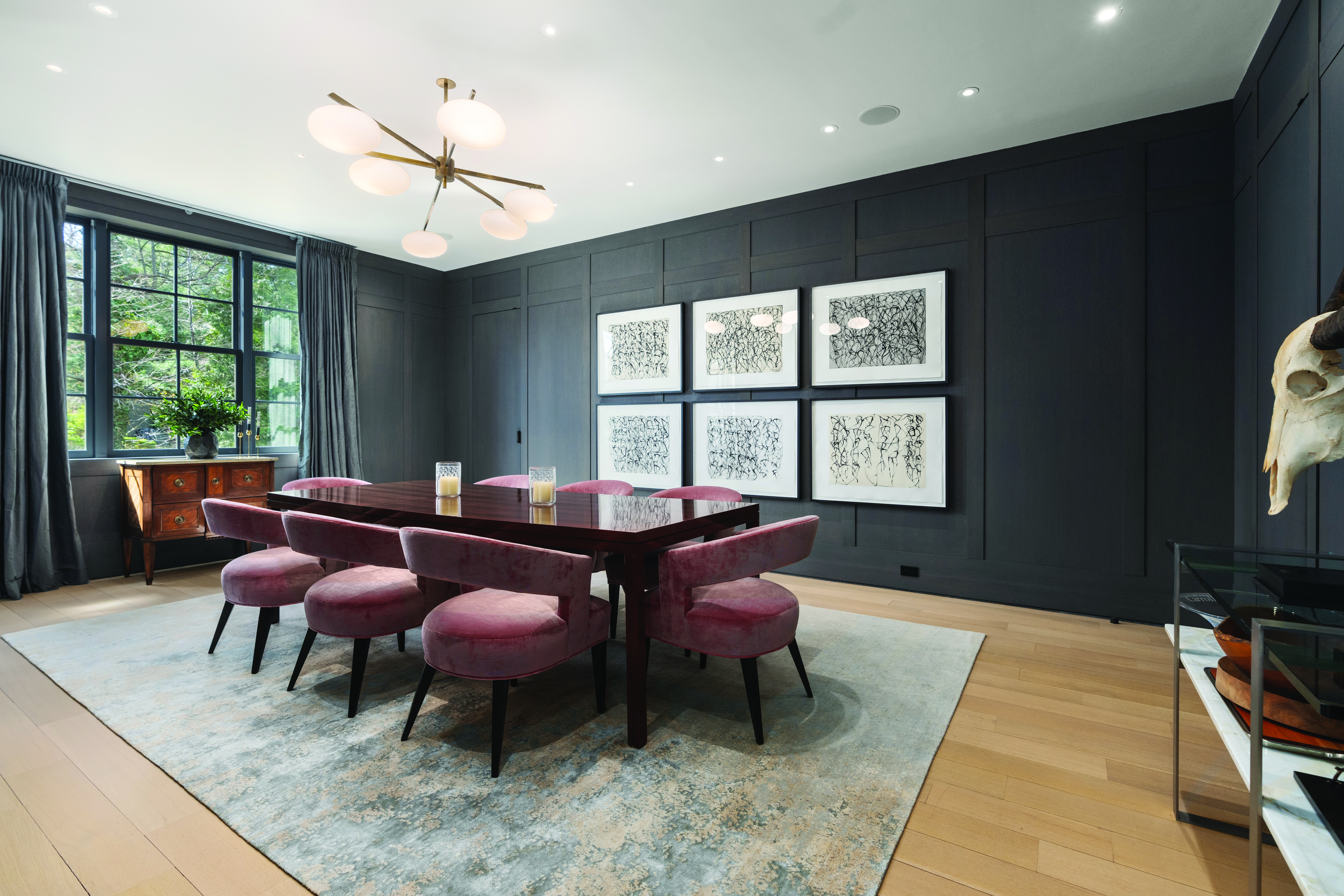 Elegant dining room featuring dark paneled walls, a modern chandelier, blush velvet chairs, and contemporary artwork in a luxury West Cambridge home.