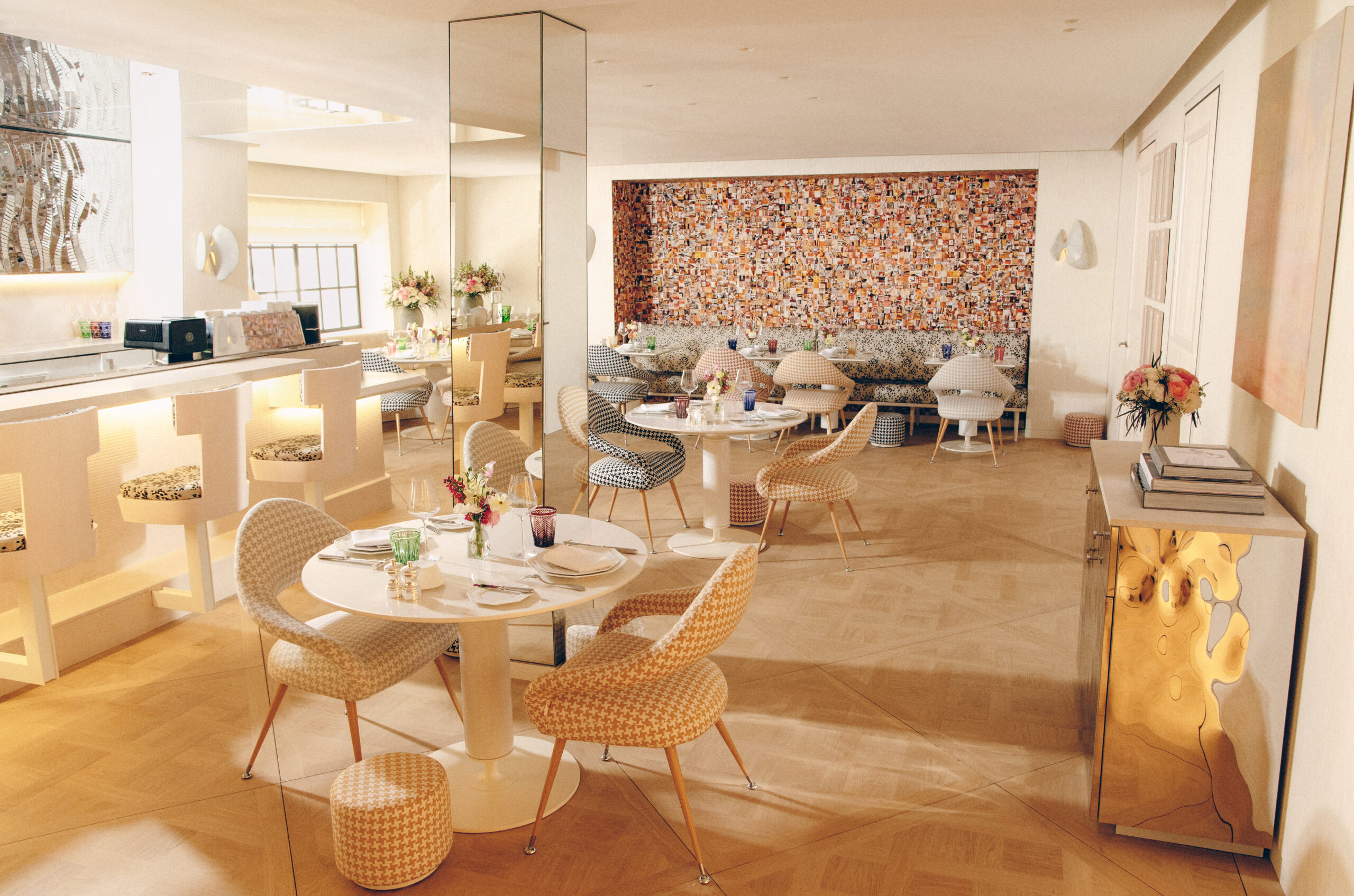 Bright, modern restaurant interior with light wood floors, round dining tables, patterned chairs, and a mosaic accent wall, featuring soft natural light and contemporary décor.
