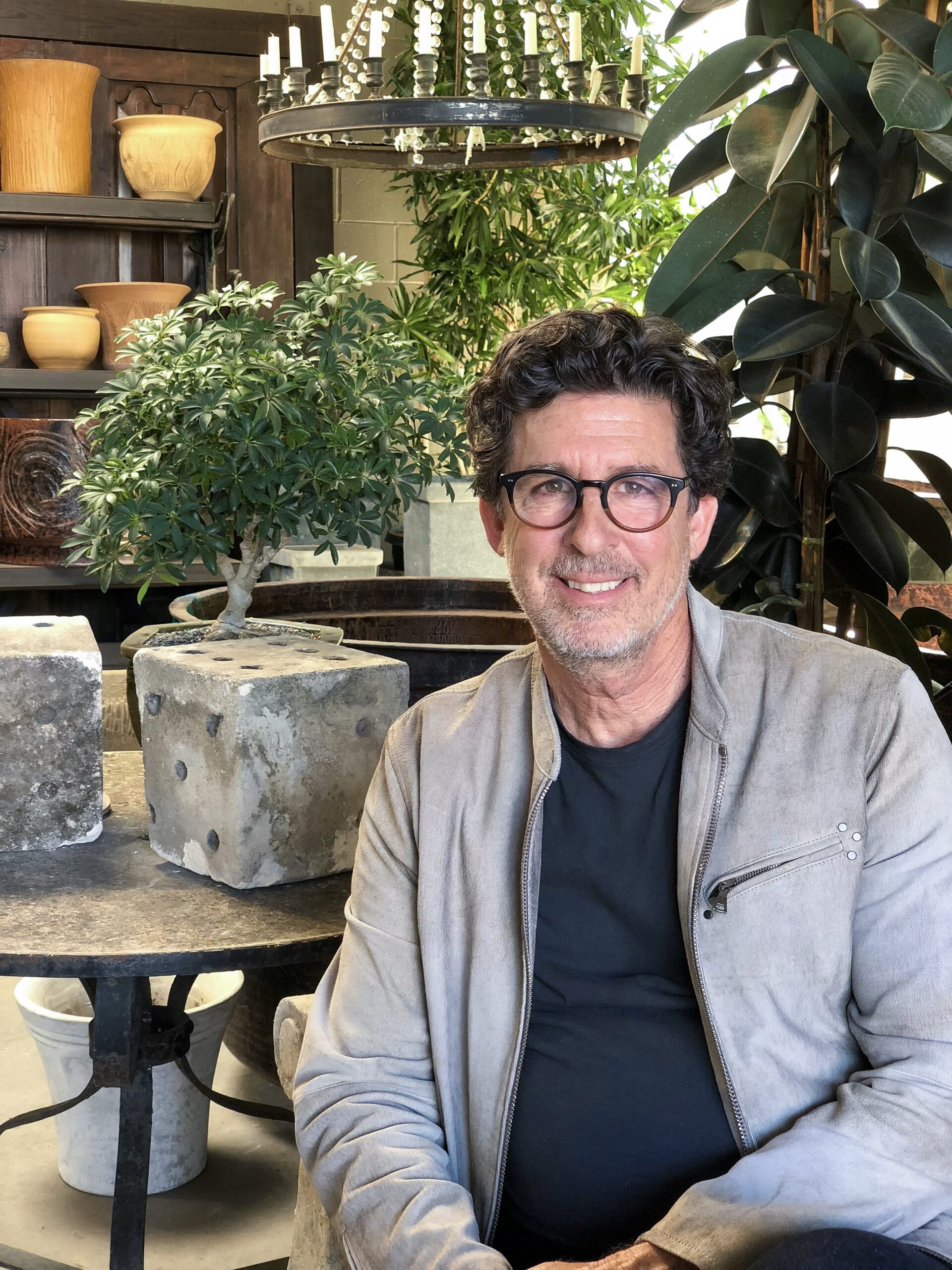 Horticulturist Stephen Block sits in a warmly lit interior filled with greenery and rustic decor. He wears glasses, a black shirt, and a light gray jacket, smiling at the camera. Behind him are potted plants, a chandelier with candles, and shelves displaying natural-toned pottery, reflecting his signature blend of organic elegance and refined design.