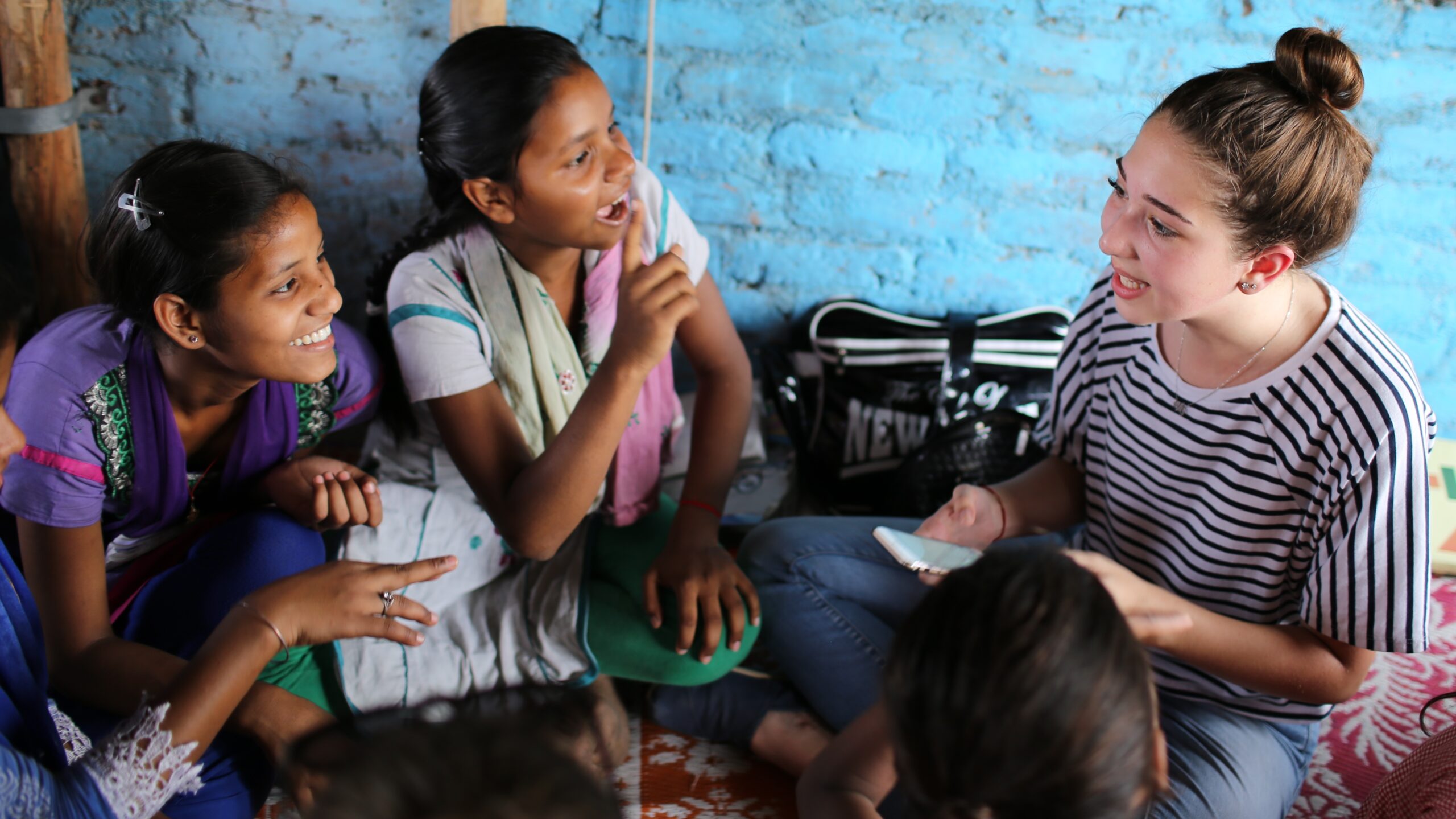 Young woman seated on the floor engaging in conversation with a small group of girls in a brightly painted room, smiling and gesturing as they interact together.